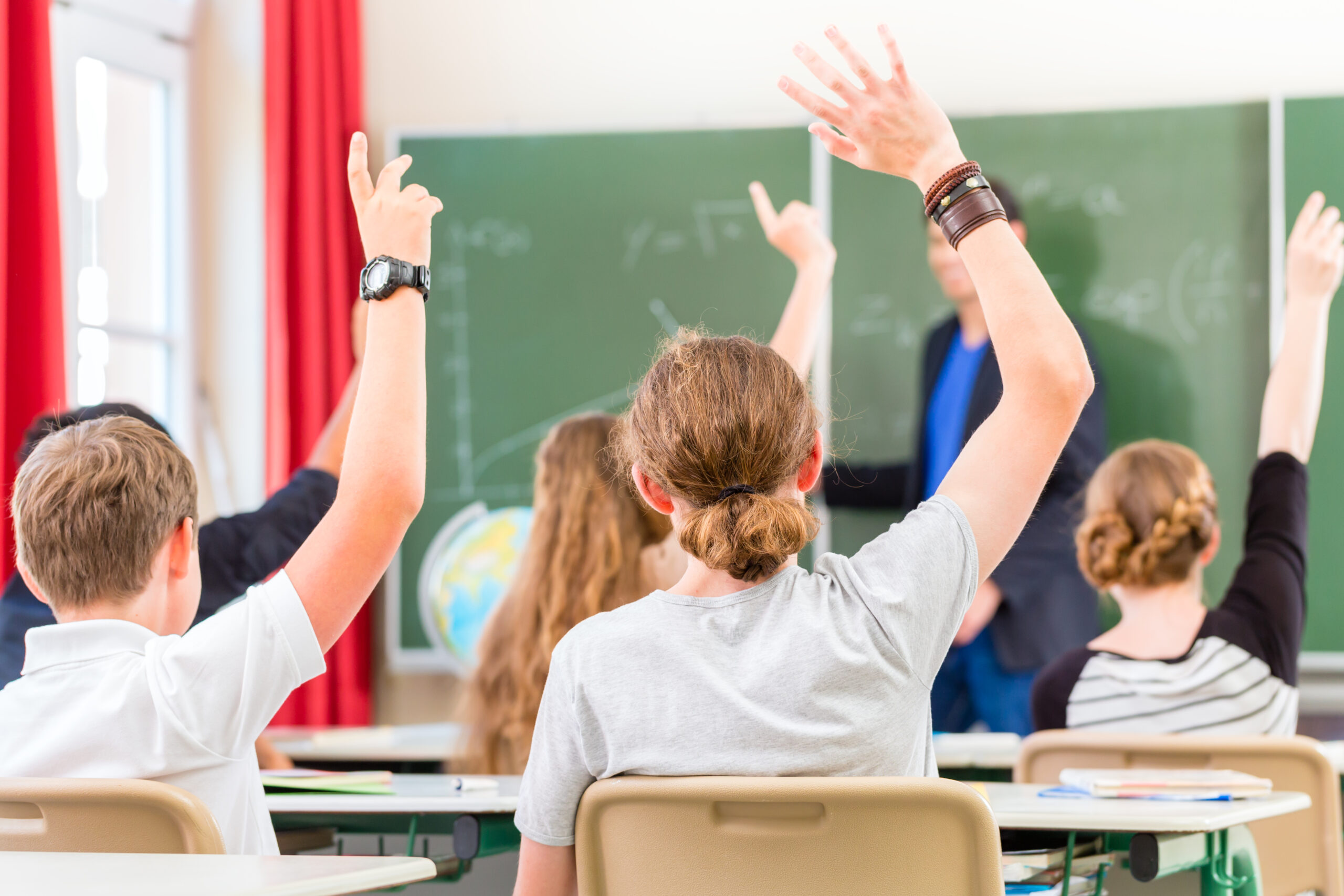 School class teacher giving lesson in front of a blackboard or board teaching students or pupils, they are raising their hands as they know all the answers