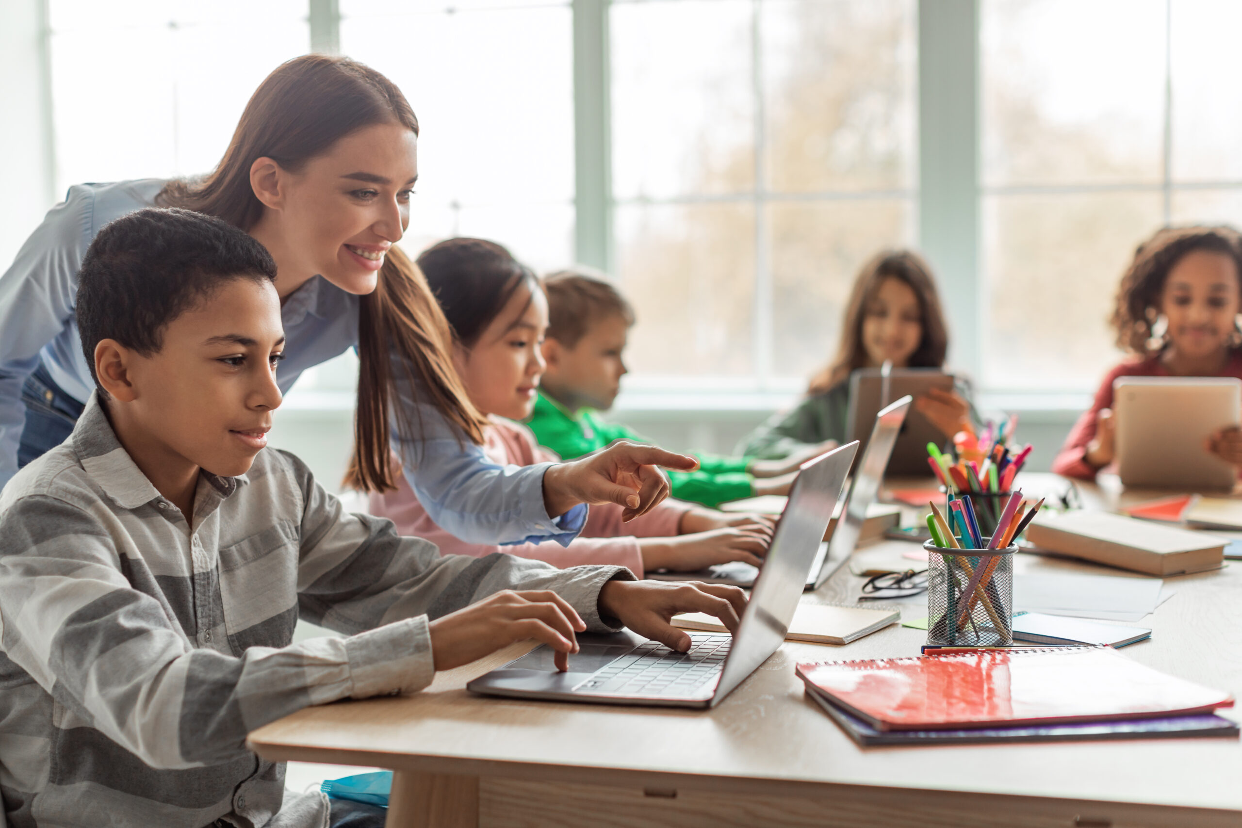 Teacher Teaching Diverse School Kids Using Laptop In Classroom. African American Schoolboy And Diverse Classmates Browsing Internet On Computer Learning Online Indoor. E-Learning Concept